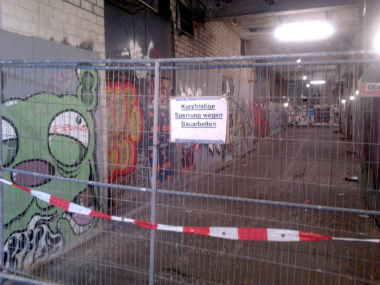 Durchgang durch das Sockelgeschoss des Ihmezentrums von der Ida-Arenhold-Brücke zur Blumenauer Straße, mit einem Baustellenzaun abgesperrt. Auf dem Zaun ein ausgedrucktes, in Plastikfolie gestecktes Stück Papier mit den Worten »Kurzfristige Sperrung wegen Bauarbeiten«.